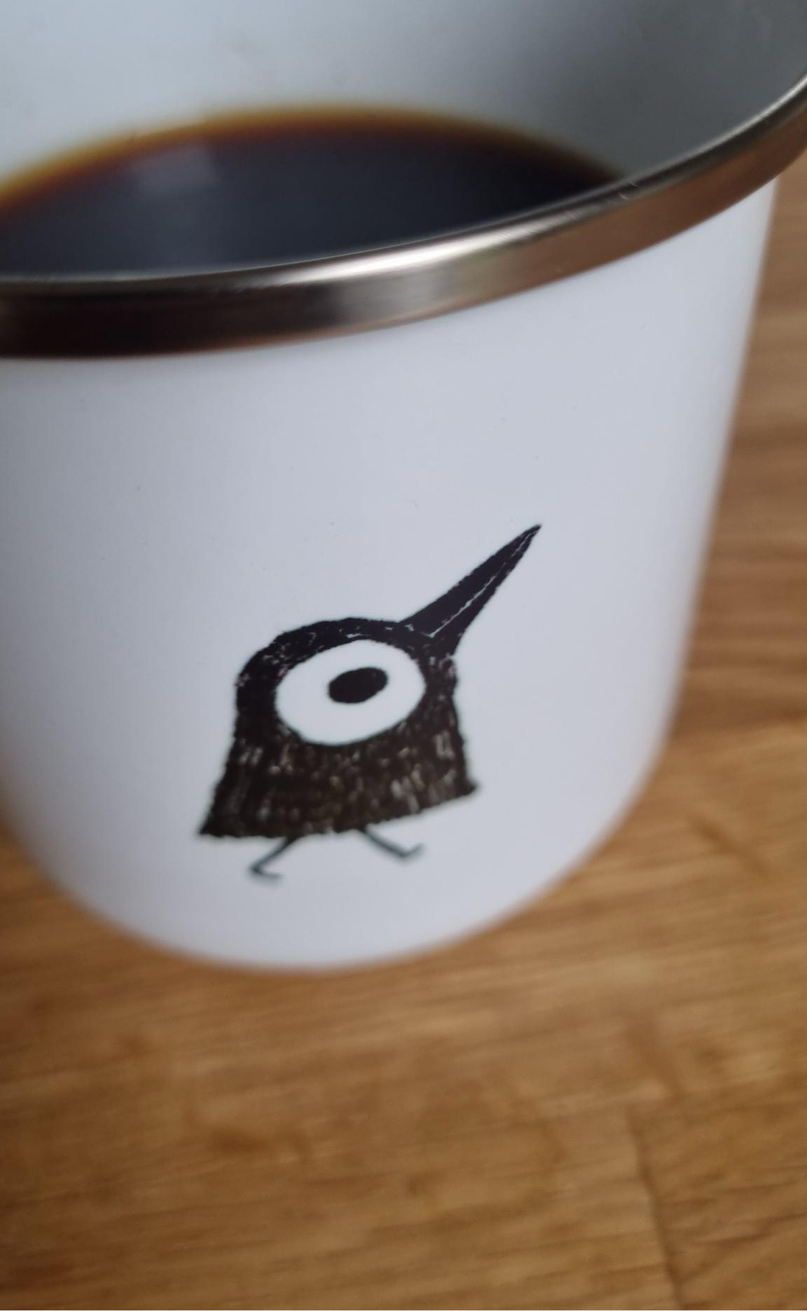Weiße mit Kaffee gefüllte Emailletasse, die mit einem kleinen schwarzen Vogel bedruckt ist. 